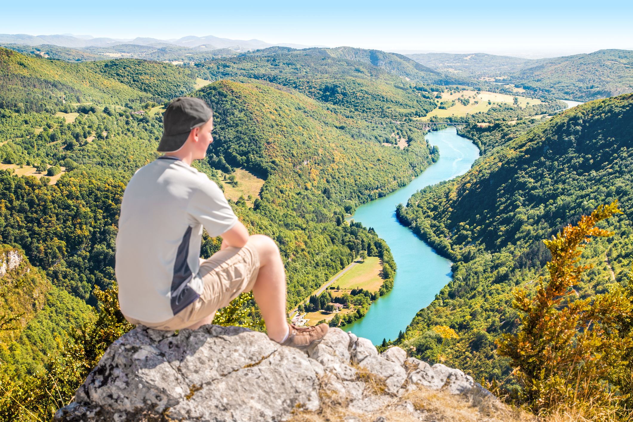 Composition en mode paysage - un jeune homme de dos, assis sur un rocher en promontoire regarde la vallée de la rivière Ain dans la région du Bugey, région Auvergne Rhône-Alpes. Photo prise par beau temps.